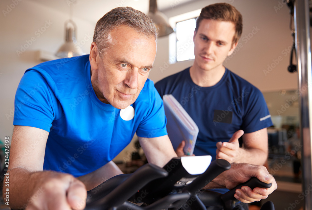Senior Man Exercising On Cycling Machine Being Encouraged By Personal Trainer In Gym