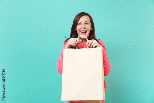Excited cheerful young woman in knitted pink sweater holding shopping bag with sale written text inscription isolated on blue turquoise wall background. People lifestyle concept. Mock up copy space.