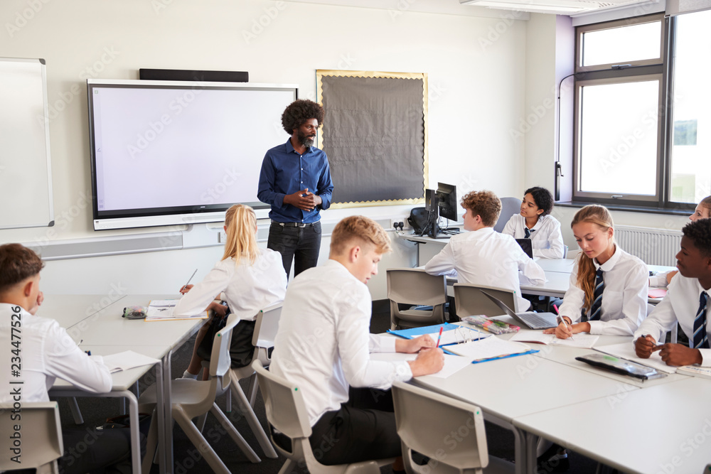 Male High School Teacher Standing Next To Interactive Whiteboard And Teaching Lesson To Pupils Wearing Uniform Male High School Teacher Standing Next To Interactive Whiteboard And Teaching Lesson To Pupils Wearing Uniform