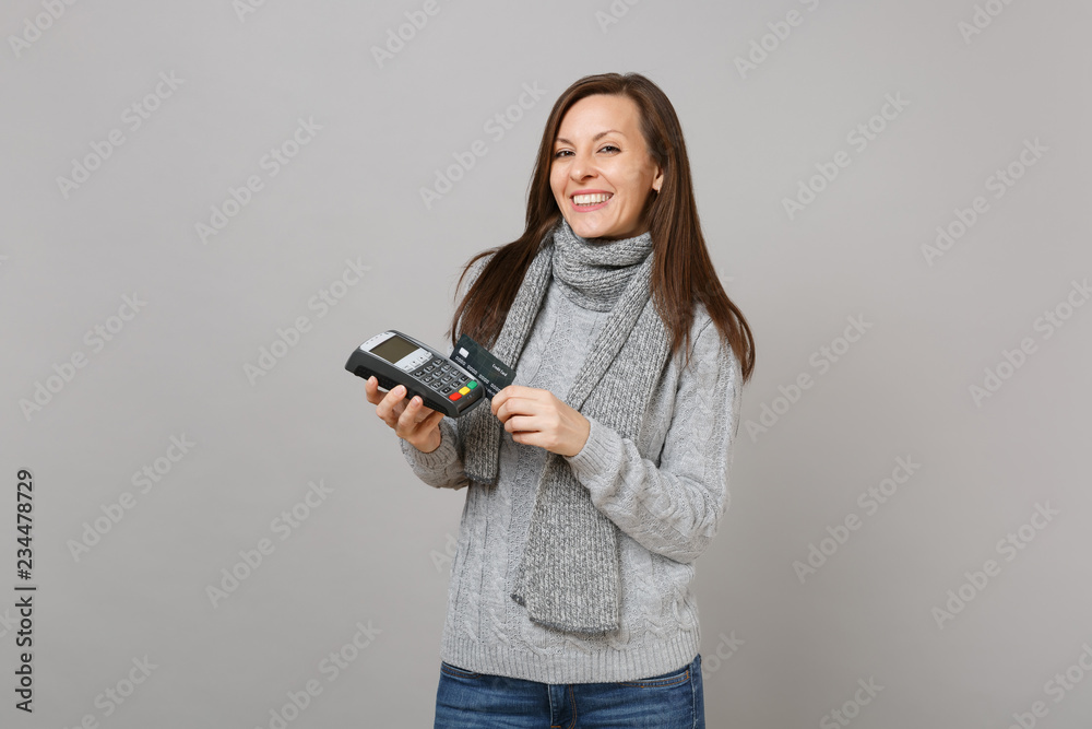 Joyful woman in sweater, scarf hold wireless modern bank payment terminal to process, acquire credit card payments isolated on grey background. Lifestyle, people sincere emotions, cold season concept. Joyful woman in sweater, scarf hold wireless modern bank payment terminal to process, acquire credit card payments isolated on grey background. Lifestyle, people sincere emotions, cold season concept.