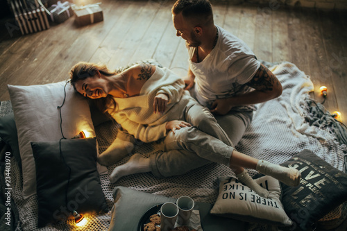 Young couple having fun and laughing cheerfully on background of coverlet, pillows and glowing lightbulbs.