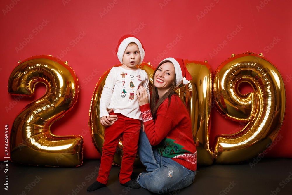 Merry Santa boy mom in Christmas hat celebrating holiday party isolated on bright red wall background golden shiny glitter numbers air balloons full length studio portrait. Happy New Year 2019 concept