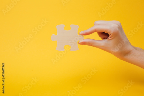 Close up cropped photo of hand holding wooden jigsaw puzzle piece isolated on bright yellow wall background. Association and connection concept. Copy space advertising mock up.