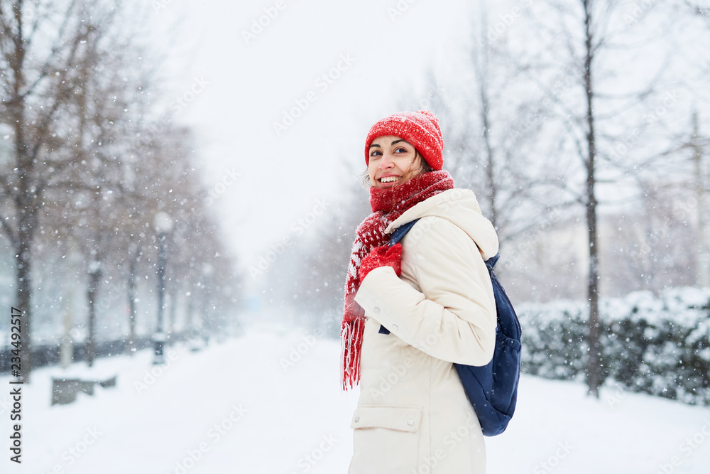 Fototapeta premium Cheerful smiling woman in white down jacket and red cap, scarf and mittens walking on the snowy street after blizzard in city. winter city after blizzards and snowfall.