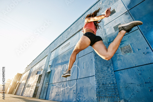 Low angle view of female athlete running along sidewalk past blue building covered in graffiti.