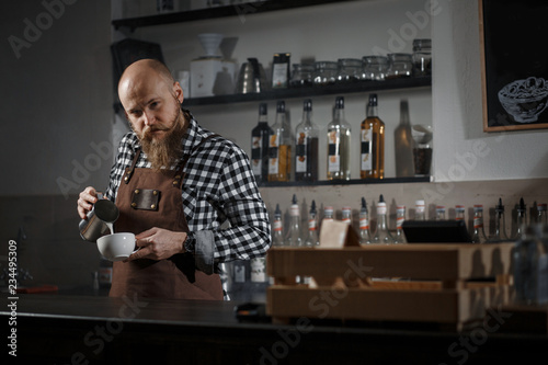 Brutal young barista in an apron makes coffee at the bar in a modern cafe