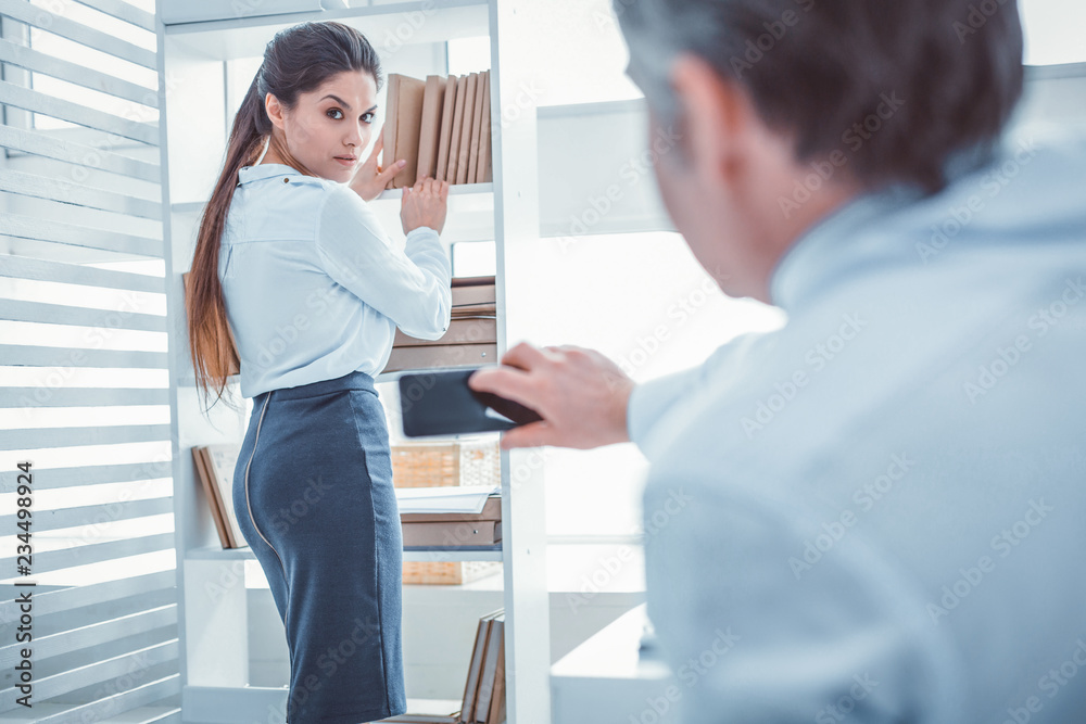 Fototapeta premium Photo without permission. Serious good-looking woman staying near shelves with documents while her workmate recording her