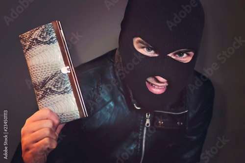 Masked criminal at the black background grins looking at the viewer and showing a purse in hand. Evil-doer is glad to have a stolen leather wallet. A satisfied robber after stealage. Thievish nature. photo