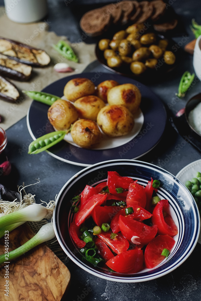 Delicious dinner table for two with fried potatoes, organic tomatoes salad, olives, green onion, eggplants on dark stone background. Delicious dinner table for two with fried potatoes, organic tomatoes salad, olives, green onion, eggplants on dark stone background.