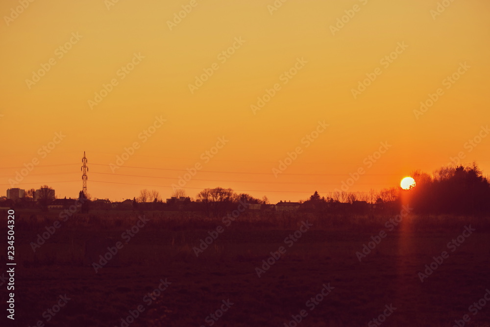 Sunset landscape, orange sky, sun setting down, dark horizon with silhouettes of electricity pylon, wires, houses and trees, rural countryside