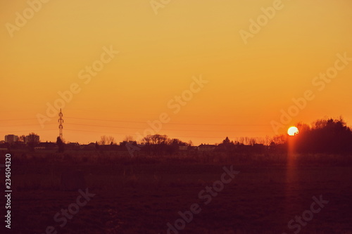 Sunset landscape, orange sky, sun setting down, dark horizon with silhouettes of electricity pylon, wires, houses and trees, rural countryside