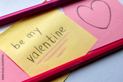 Be My Valentine handwritten inscription on the yellow sticker and the heart drawn on a pink paper with two pencils