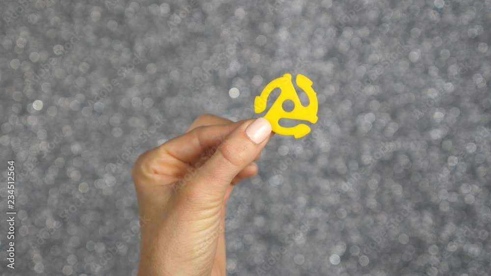Female hand holds yellow 45 rpm record adaptor up. This is a small insert that goes in the middle of a 45 rpm record so it can be played on the spindle of a record player. Silver background. Female hand holds yellow 45 rpm record adaptor up. This is a small insert that goes in the middle of a 45 rpm record so it can be played on the spindle of a record player. Silver background.