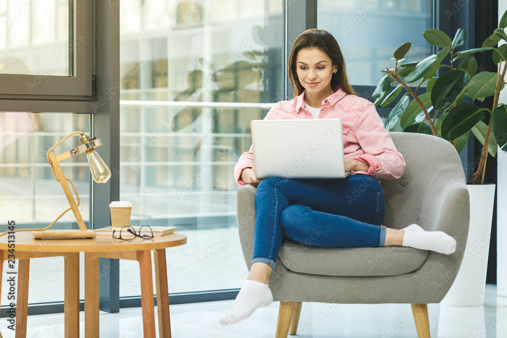 Enjoying working time at home. Beautiful young smiling casual woman working on laptop and drinking coffee while sitting in a big comfortable chair at home. Enjoying working time at home. Beautiful young smiling casual woman working on laptop and drinking coffee while sitting in a big comfortable chair at home.