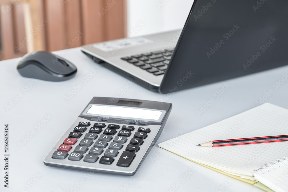 Calculator, mouse, computer, laptop and pencil are on the notebook. Placed on a white desk. Calculator, mouse, computer, laptop and pencil are on the notebook. Placed on a white desk.