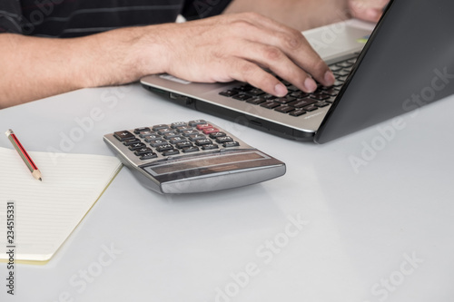 A calculator placed next to a notebook computer with a working man's hand. On a white desk and with a pencil on a notebook.