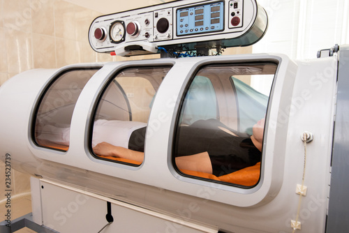 Treatment in an oxygen capsule. A beautiful girl in a black T-shirt and white pants is lying in a pressure chamber. The concept of modern medicine