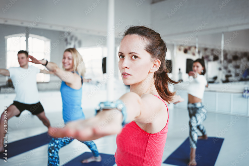 Obraz premium Portrait of handsome woman practice yoga exercises indoor loft gym with group people. Slim girl doing warrior of light asana. Blurred background