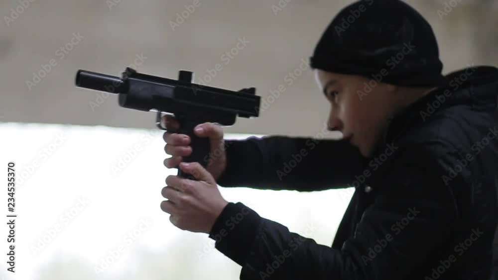 Focused Caucasian boy in black clothes holding gun in hands, looking straight. Teen pointing. Abandoned building. Indoors, autumn. Dangerous game. Focused Caucasian boy in black clothes holding gun in hands, looking straight. Teen pointing. Abandoned building. Indoors, autumn. Dangerous game.