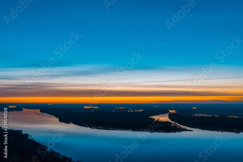 Beautiful river with big island with trees under predawn sky. Bright orange stripe in picturesque cloudy sky. Early blue sky reflected in water. Colorful morning atmospheric image of majestic nature.