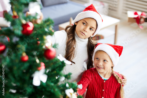 Two little girls in Santa caps looking at decorations on Christmas tree while standing near by