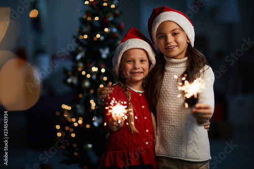 Cheerful little girls in xmas attire holding burning Bengal lights on Christmas night