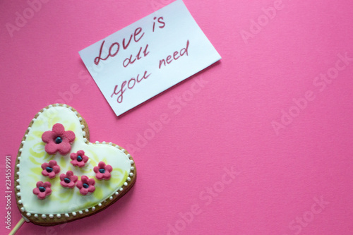 Gingerbread heart with white icing and pink flowers shit of paper on pink background. Love concept. Gift for womans and valentines day. Copy space for text.