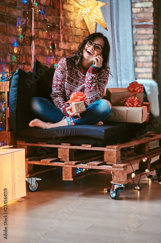 Cheerful beautiful girl talking by phone with friends while sitting on a couch with gift boxes in a decorated room with loft interior.