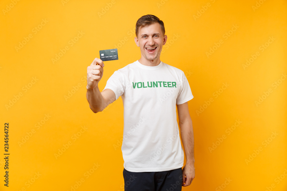 Portrait of man in white t-shirt with written inscription green title volunteer hold in hand credit bank card isolated on yellow background. Voluntary free assistance help, charity grace work concept. Portrait of man in white t-shirt with written inscription green title volunteer hold in hand credit bank card isolated on yellow background. Voluntary free assistance help, charity grace work concept.