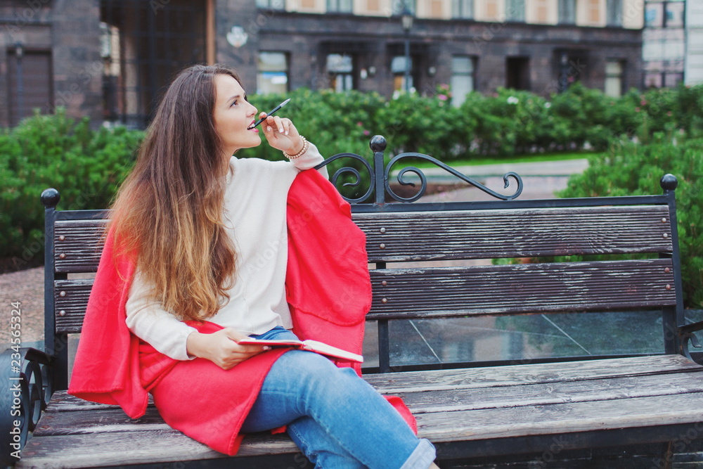 An attractive girl with long brown hair sits on a bench, hiding behind a red rug, gnawing a pen and thinking on an urban background. She has a notebook in her hands An attractive girl with long brown hair sits on a bench, hiding behind a red rug, gnawing a pen and thinking on an urban background. She has a notebook in her hands