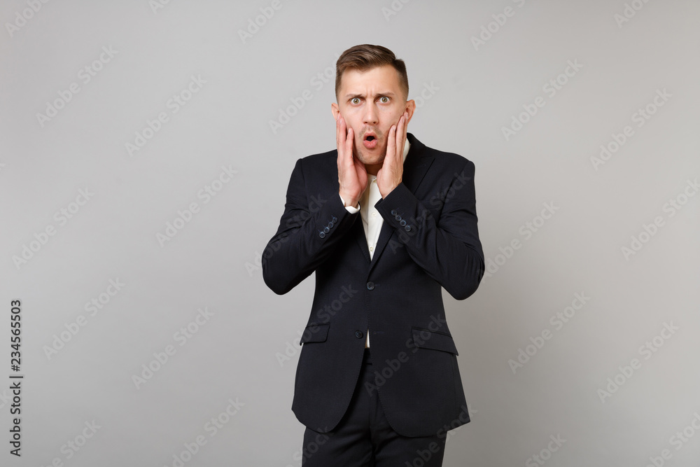 Portrait of shocked young business man in classic black suit, shirt putting hands on cheeks isolated on grey wall background in studio. Achievement career wealth business concept. Mock up copy space. Portrait of shocked young business man in classic black suit, shirt putting hands on cheeks isolated on grey wall background in studio. Achievement career wealth business concept. Mock up copy space.