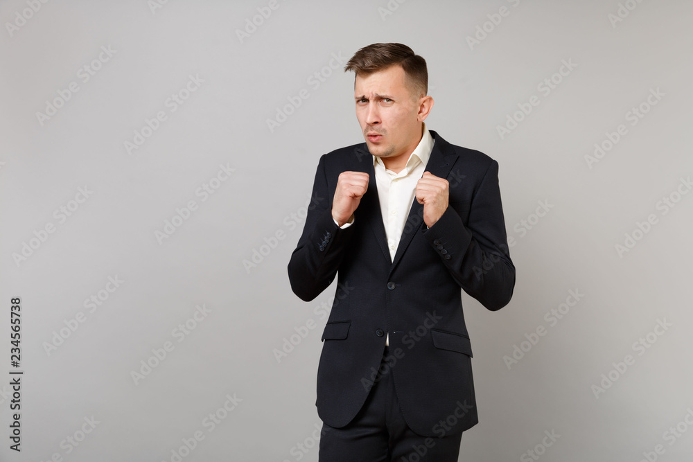 Portrait of worried scared young business man in classic black suit, shirt clenching fists isolated on grey wall background in studio. Achievement career wealth business concept. Mock up copy space. Portrait of worried scared young business man in classic black suit, shirt clenching fists isolated on grey wall background in studio. Achievement career wealth business concept. Mock up copy space.
