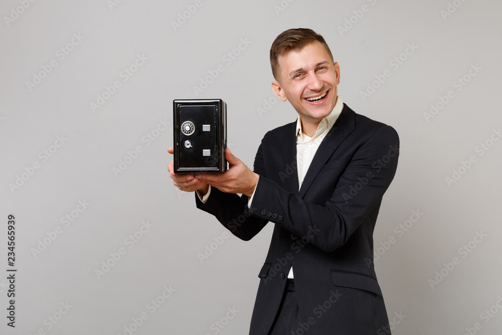 Cheerful young business man in classic black suit and shirt holding metal bank safe for money accumulation isolated on grey background. Achievement career wealth business concept. Mock up copy space.