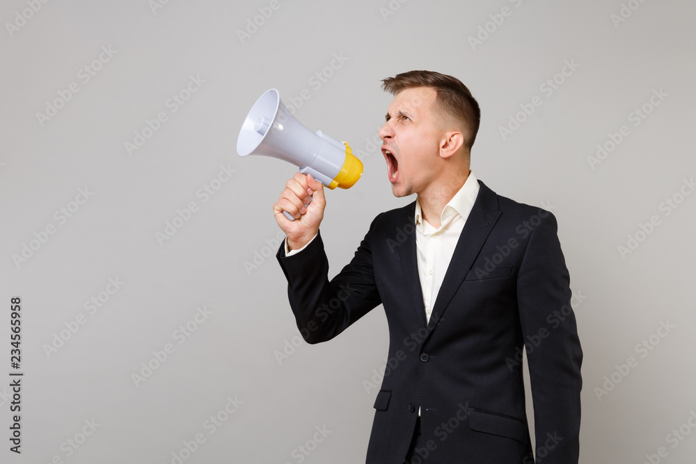 Irritated young business man in classic black suit, shirt looking aside, screaming on megaphone isolated on grey background in studio. Achievement career wealth business concept. Mock up copy space. Irritated young business man in classic black suit, shirt looking aside, screaming on megaphone isolated on grey background in studio. Achievement career wealth business concept. Mock up copy space.