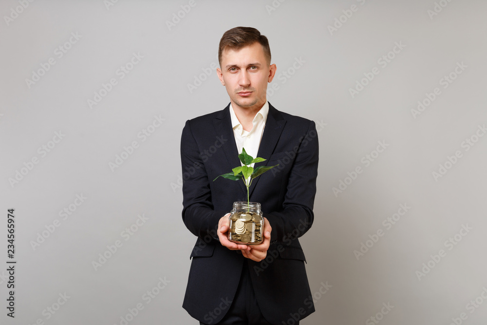 Young business man in classic black suit shirt holding pile gold coins in glass jar with green plant sprout isolated on grey background. Achievement career wealth business concept. Mock up copy space. Young business man in classic black suit shirt holding pile gold coins in glass jar with green plant sprout isolated on grey background. Achievement career wealth business concept. Mock up copy space.