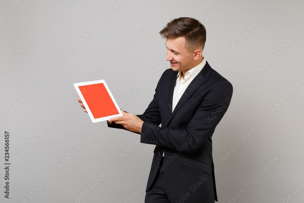 Young business man in classic black suit holding and looking on tablet pc computer with blank empty screen isolated on grey background. Achievement career wealth business concept. Mock up copy space. Young business man in classic black suit holding and looking on tablet pc computer with blank empty screen isolated on grey background. Achievement career wealth business concept. Mock up copy space.
