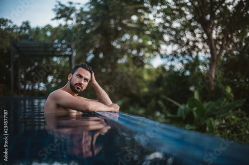Portrait of a young handsome man in an outdoor pool in the tropics. Attractive guy with a beard at the side of the pool © Anna