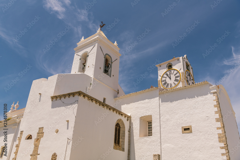 White, bell tower, clock tower, and walls of the historic church of Igreja de Santa Maria do Castelo in Tavira, Portugal in brilliant sunlight under a blue sky