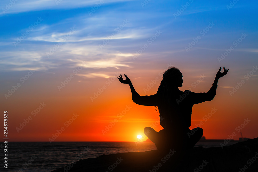 Yoga silhouette of woman meditating on the ocean beach during amazing sunset.