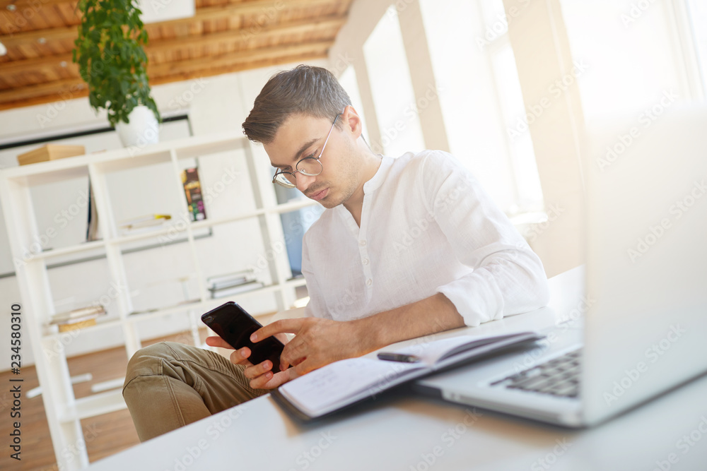 Fototapeta premium Portrait of pensive serious young businessman wears white shirt and glasses working with laptop and using mobile phone at the table in office