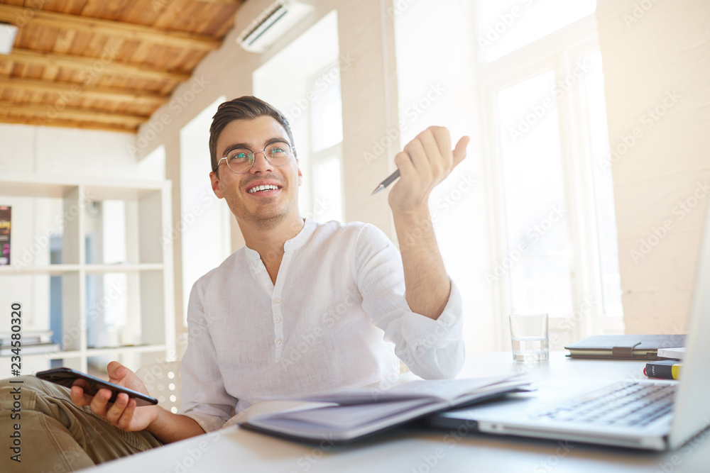 Closeup of smiling attractive young businessman wears white shirt and spectacles using smartphone and writing in notebook at the table in office Closeup of smiling attractive young businessman wears white shirt and spectacles using smartphone and writing in notebook at the table in office