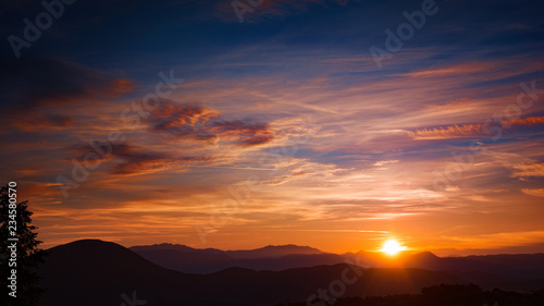 sun setting behind silhouetted mountains in a blue sky with travelling clouds © christos