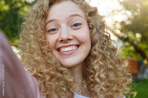 Young beautiful funny cool girl blonde with curly hair, widely smiling and looking into the camera, making selfie, nature background photo