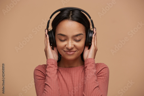 Portrait of a young mixedrace girl listens to her favorite music in big black headphones, closing her eyes from pleasure, enjoyment, music lover, likes lyric songs about love, on beige background