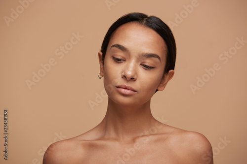 Beautiful caucasian girl, looking down, after a skincare and spa, with healthy clean fresh skin. Pretty woman with black hair fixed behind, thick eyebrows and naked shoulders over beige background photo