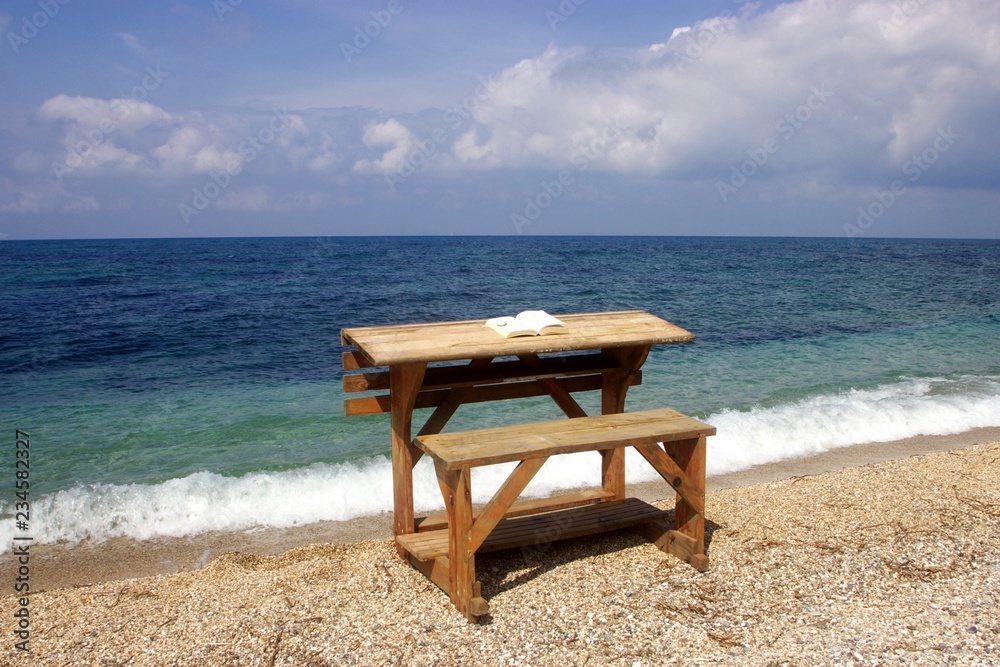 Wooden desk on a beach with an open book on it and blue sea and clouds at the background 