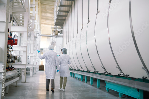 Back view full length portrait of two factory workers wearing white coats crossing production workshop, copy space photo