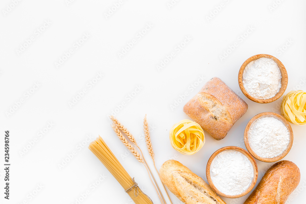 Products made of wheat flour. White flour in bowl, wheat ears, fresh bread and raw pasta on white background top view space for text Products made of wheat flour. White flour in bowl, wheat ears, fresh bread and raw pasta on white background top view space for text