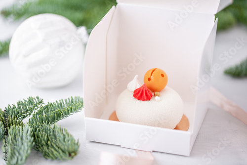 Mini mousse pastry dessert covered with white velour. Individually wrapped in white box. The branches of spruce on bokeh background. Modern european cake. French cuisine. Christmas . Snowflake