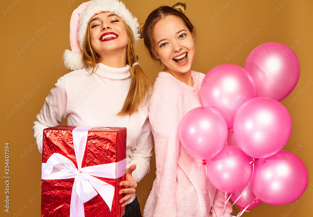 Christmas,x-mas, concept.Two smiling beautiful women in white winter sweaters.Girls posing on golden background.Models with big gift box and pink balloons.Having fun,ready for celebration new year Christmas,x-mas, concept.Two smiling beautiful women in white winter sweaters.Girls posing on golden background.Models with big gift box and pink balloons.Having fun,ready for celebration new year